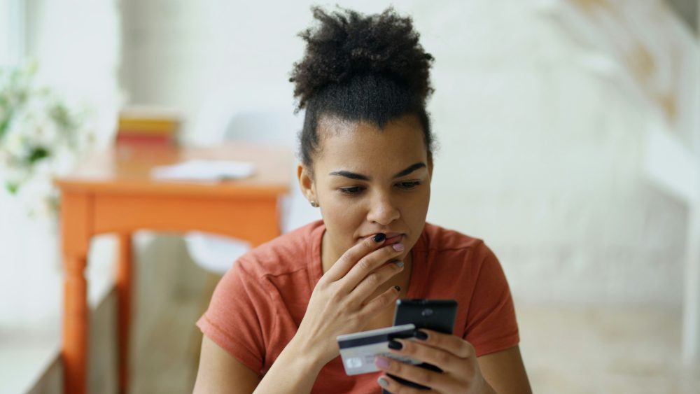 A woman in an orange t-shirt sits at a table looking at a credit card, with worry on her face.