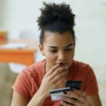 A woman in an orange t-shirt sits at a table looking at a credit card, with worry on her face.