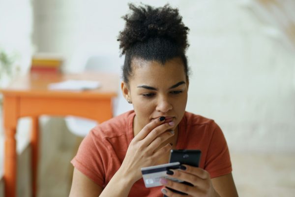 A woman in an orange t-shirt sits at a table looking at a credit card, with worry on her face.